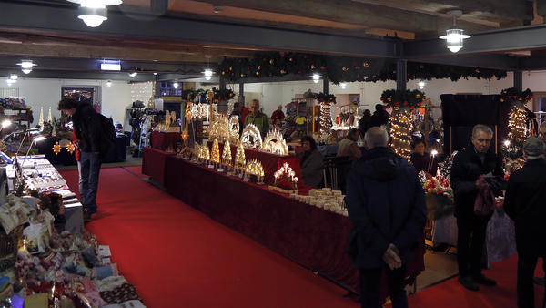 In einem Raum stehen mehrere Menschen an Verkaufstischen, auf denen verschiedene, insbesondere weihnachtliche, Dekorationsartikel angeboten werden.