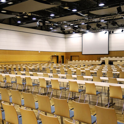 Zu Sehen ist eine parlamentarische Bestuhlung aus Stühlen und Tischen sowie eine Reihenbestuhlung, eine Bühne mit Podiumsplätzen, eine große Leinwand sowie Veranstaltungstechnik.