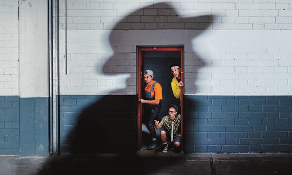 Auf dem Foto sind drei Personen zu sehen, die aus einem Türrahmen herausschauen. Im Hintergrund ist zudem ein großer Schatten eines Mannes mit Hut zu sehen.